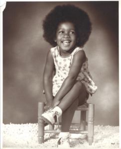 Young toddler Angela with an afro sitting on a wooden chair wearing a white dress and white shoes