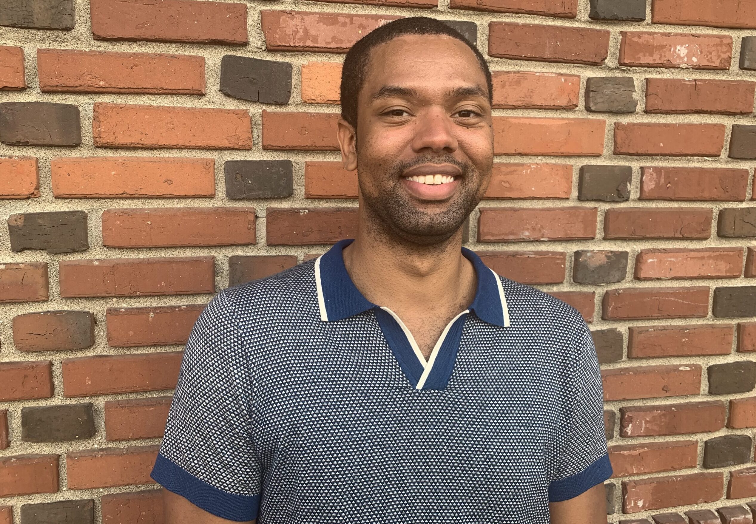 Tremayne standing in front of a brick wall, wearing a blue collared shirt