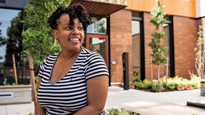 A black woman sits outside a modern building, smiling and looking to the side.