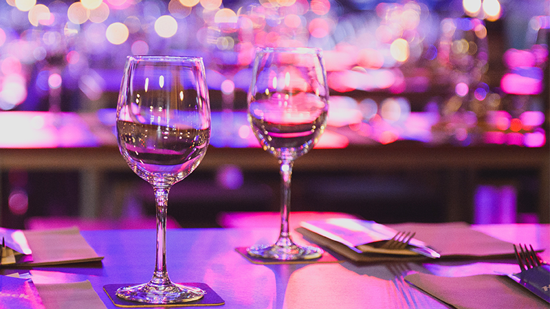 Two wine glasses on a table with place settings and bright lights