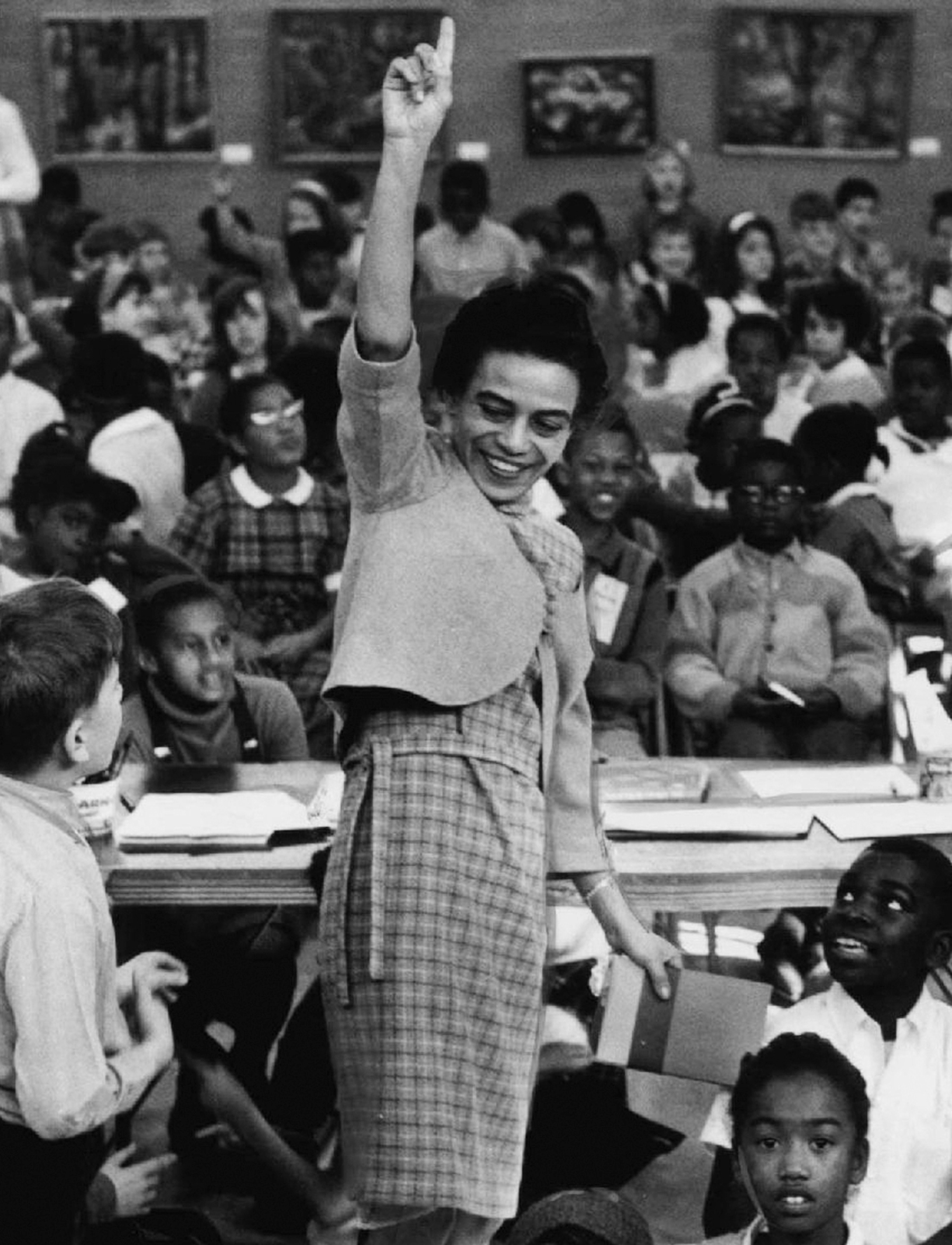 A black and white photograph of a woman, surrounded by school children, pointing her finger toward the sky.