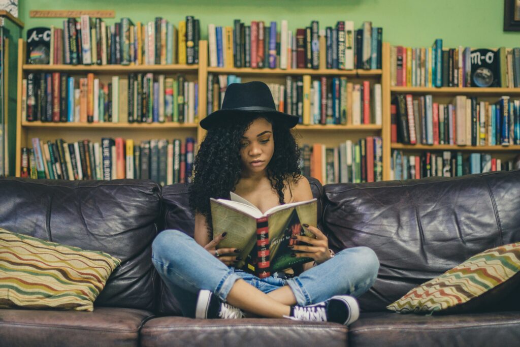 A black women wearing a hat sitting on a couch in a library reading a book