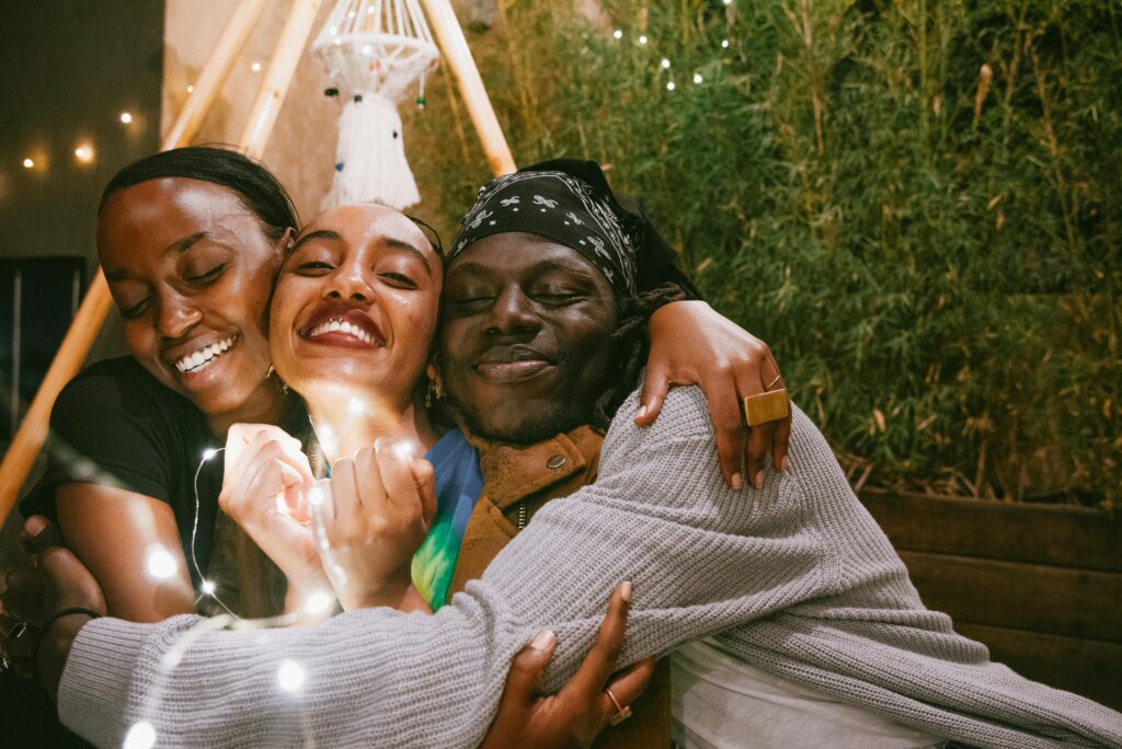 Three black friends hugging each other with smiles on their faces, outdoors under twinkling lights