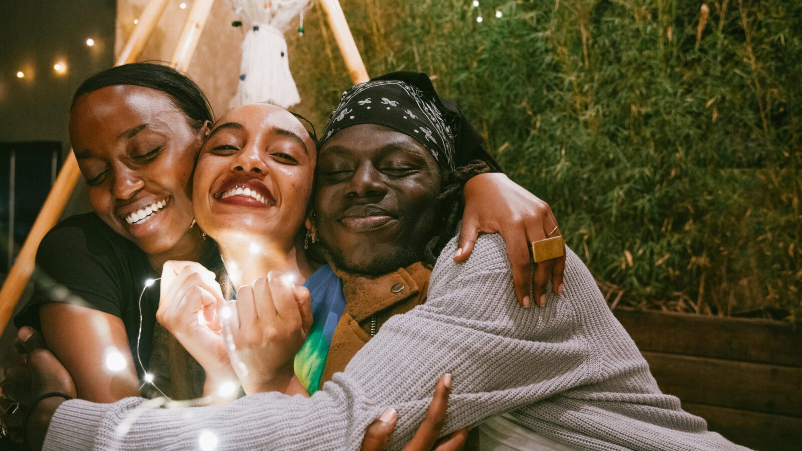 Three black friends hugging each other with smiles on their faces, outdoors under twinkling lights