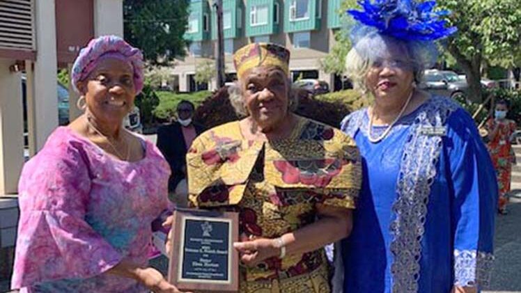 Ms. Elma Horton stands between two other women — all wearing dresses and hats — holding up a plaque.