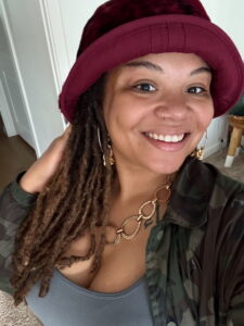 Headshot of a woman with brown hair, wearing a red hat, green shirt, and gold necklace